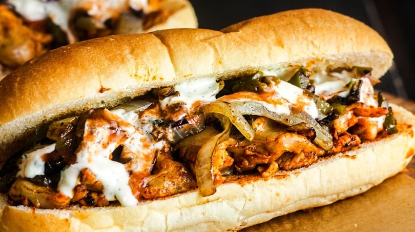 Close-up of a chicken cheesesteak sandwich with melted cheese, peppers, onions, and sauce on a bun.