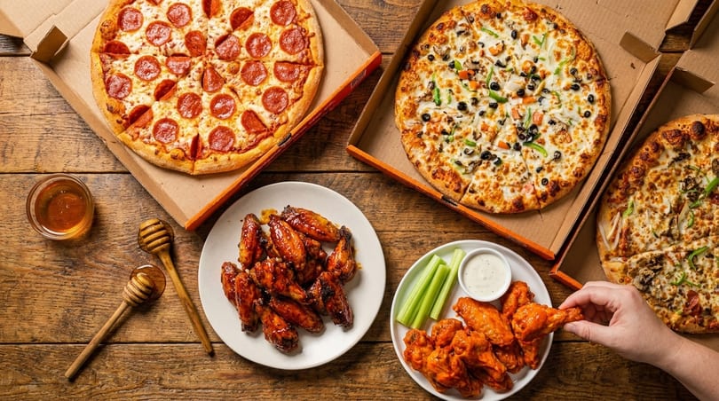 Three pizzas, two plates of chicken wings, and honey on a rustic wooden table.