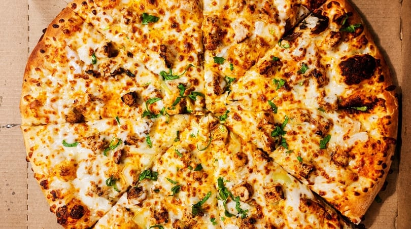 Close-up of a whole chicken pizza with melted cheese and green herbs in a cardboard box.